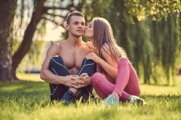 Έννοια του υγιεινού τρόπου ζωής. Χαρούμενος νεαρός γυμναστήριο ζευγάρι φιλί, ενώ κάθεται σε ένα πράσινο γρασίδι. — Φωτογραφία Αρχείου