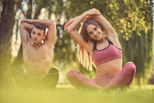 Koncept zdravého životního stylu. Pár šťastný mladý fitness strečink ruce, zatímco sedí v lotosu představují na zelené trávě. — Stock fotografie