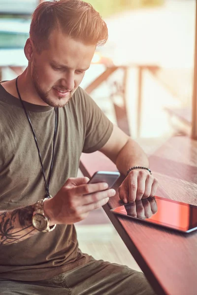 Knappe stijlvolle hipster zit aan een tafel in een café langs de weg, het lezen van het bericht op de smartphone. — Stockfoto