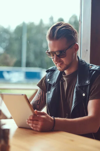 Hipster chipeș cu o tunsoare elegantă și barbă stă la o masă într-o cafenea pe marginea drumului, arată ceva în computerul tabletei . — Fotografie, imagine de stoc