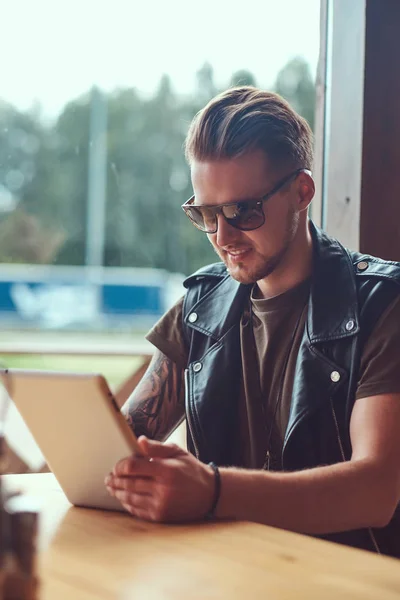 Όμορφος hipster με μοντέρνο κούρεμα και γενειάδα κάθεται σε ένα τραπέζι σε ένα καφέ άκρη του δρόμου, μοιάζει κάτι στον υπολογιστή tablet. — Φωτογραφία Αρχείου