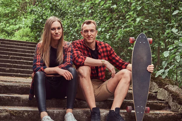 Junges Hipster-Paar, gutaussehender Mann mit Skateboard und blondem Mädchen, sitzt auf Stufen in einem Park. — Stockfoto