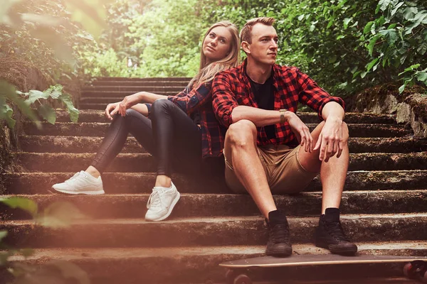 Pár mladých bokovky, pohledný muž s skateboard a blondýna dívka, sedící kroky v parku. — Stock fotografie