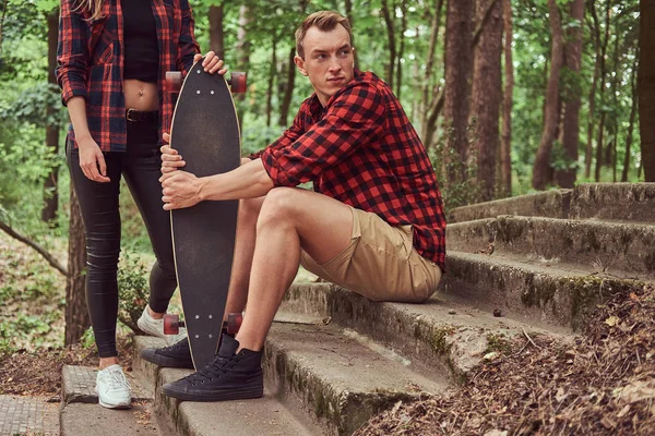 Giovane coppia hipster, bell'uomo con uno skateboard e una ragazza bionda, seduta su dei gradini in un parco . — Foto Stock