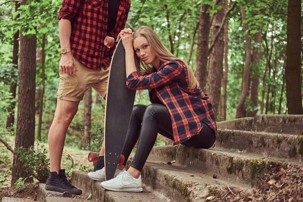 Giovane coppia hipster, bell'uomo e ragazza bionda con uno skateboard, seduta su gradini in un parco . — Foto Stock