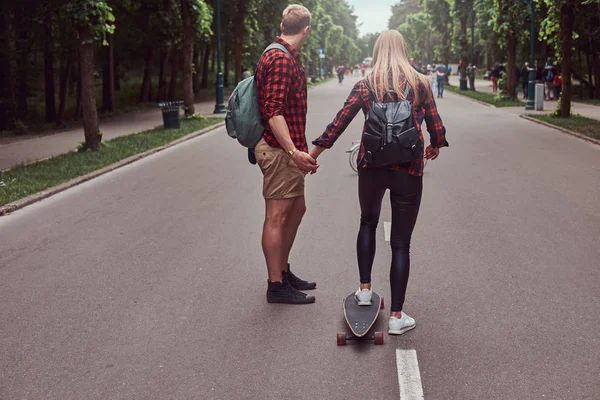 Giovane coppia hipster. Guy insegna alla sua ragazza a pattinare per strada in un parco . — Foto Stock