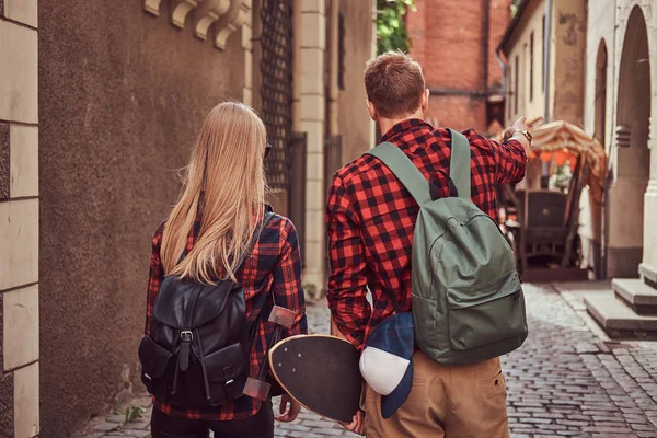 Vue arrière d'un jeune couple hipster, beau patineur et sa petite amie, se tenant la main, se promenant dans une vieille rue étroite d'Europe . — Photo