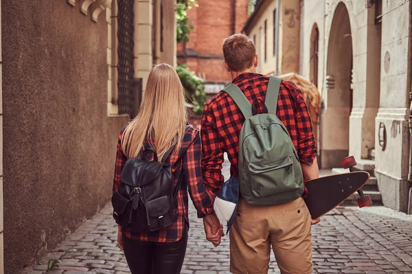 Baksidan på en ung hipster par, stilig skater och hans flickvän, hålla händer, gå runt en gamla smala gatorna i Europa. — Stockfoto