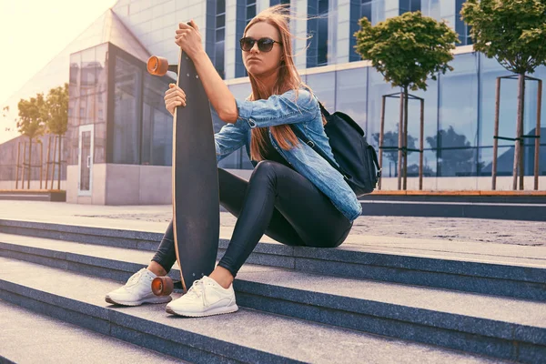 Mladí bokovky blondýnku v neformálním oblečení a sluneční brýle sedí na kroky proti mrakodrapu, odpočívá po jízdě na skateboardu. — Stock fotografie