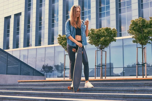 Mladí bokovky blondýnka v neformálním oblečení, stojící na kroky proti mrakodrapu, pomocí smartphone, odpočívá po jízdě na skateboardu. — Stock fotografie