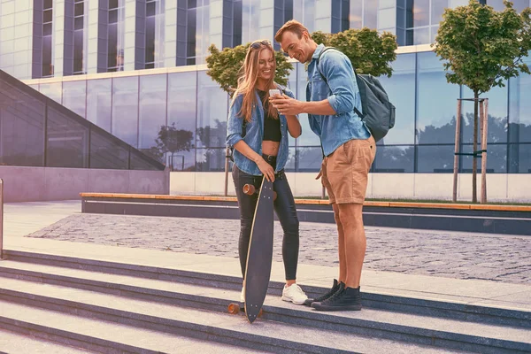 Joven pareja atractiva feliz en ropa casual de pie contra un rascacielos, viendo algo en un teléfono inteligente . — Foto de Stock