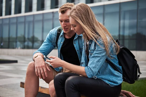 Joven feliz pareja atractiva en ropa casual sentado en un banco contra un rascacielos, viendo algo en un teléfono inteligente . — Foto de Stock