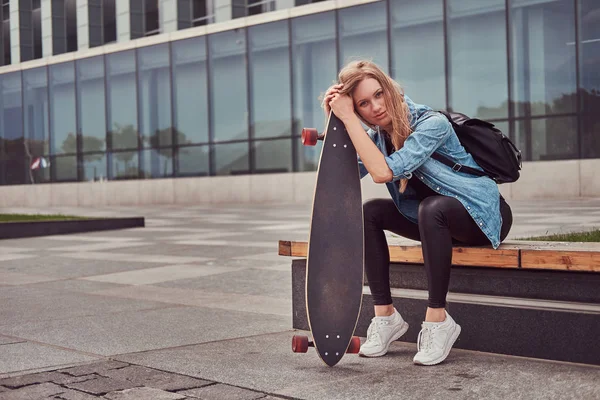 Giovane ragazza bionda hipster in abbigliamento casual seduto su una panchina contro un grattacielo, a riposo dopo aver cavalcato sullo skateboard . — Foto Stock