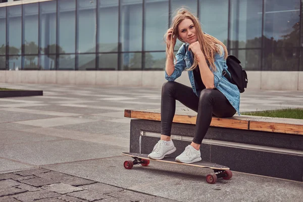 Jonge hipster blond meisje in casual clothessitting op een bank tegen een wolkenkrabber, rust na het rijden op skateboard. — Stockfoto