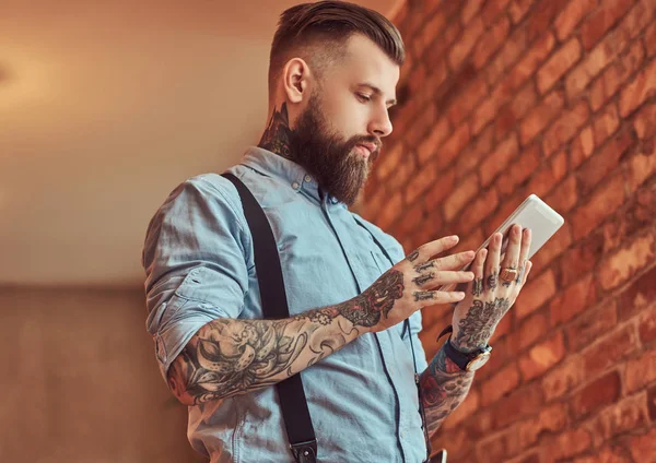 Old-fashioned tattooed hipster wearing a shirt and suspenders, in a sunglasses, using a tablet while standing near a desk in an office with loft interior.