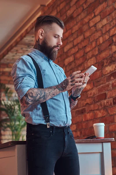 Hipster tatuado a la antigua con una camisa y tirantes, en gafas de sol, usando una tableta mientras está de pie cerca de un escritorio en una oficina con interior loft . —  Fotos de Stock