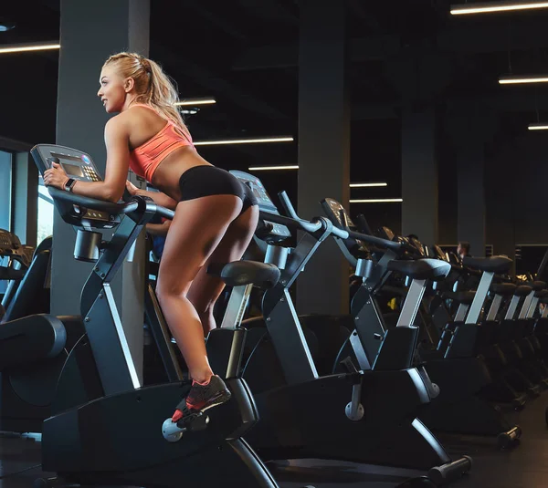 Mulher loira bonita nova no treinamento de sportswear em uma bicicleta de exercício no centro de fitness moderno . — Fotografia de Stock