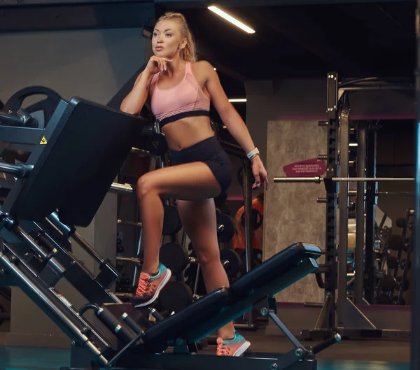 Beautifull fitness kvinna i sportkläder posing nära av ben pressmaskin i ett gym. — Stockfoto