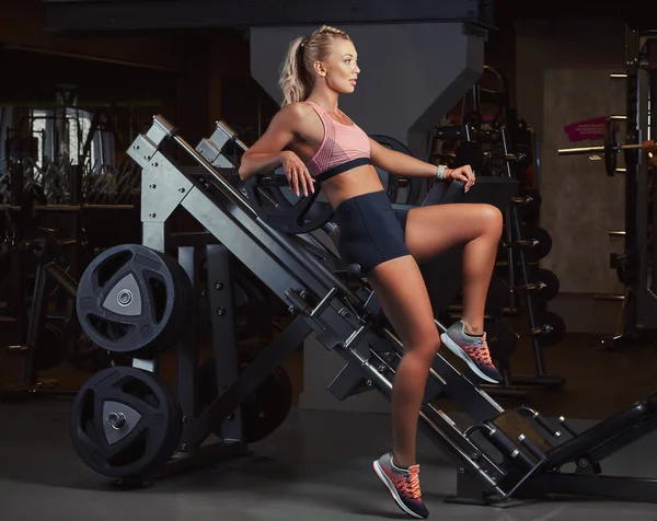 Atletisk blond kvinna i sportkläder liggande på en ben pressmaskin i gymmet. — Stockfoto