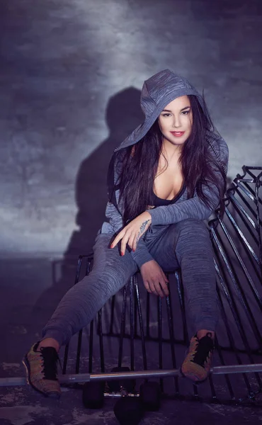 Portrait of a cool seductive brunette girl in a sexy sports bra and gray hoodie sitting on a chair in a studio. — Stock Photo, Image