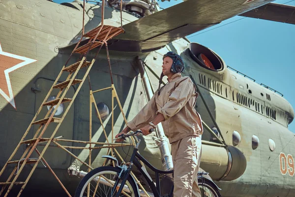 Mechanic in uniform and flying helmet walking with a bicycle near the large military helicopter in an open-air museum. — Stock Photo, Image