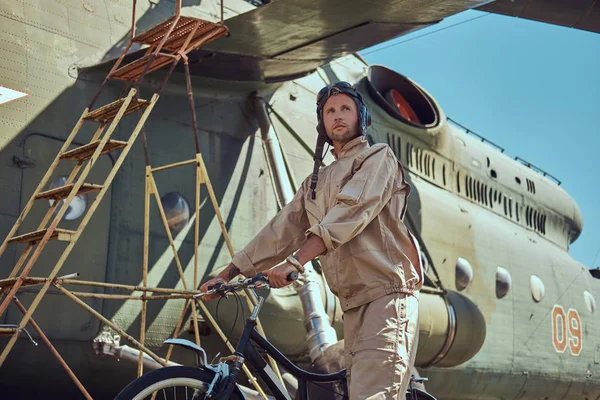 Mechanic in uniform and flying helmet walking with a bicycle near the large military helicopter in an open-air museum. — Stock Photo, Image