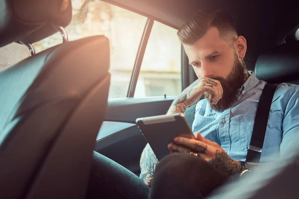 Thoughtful old-fashioned tattooed hipster guy in a shirt with suspenders, using a tablet while sitting in a luxury car on back seat. — Stock Photo, Image