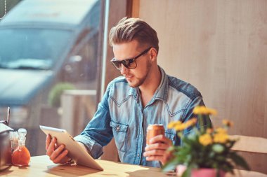 Bir şık saç ve sakal hipster adam yol kenarına yerleştirilen bir kafede bir masada oturuyor, gazoz görünüyor süre bir şey tablet içecekler. 