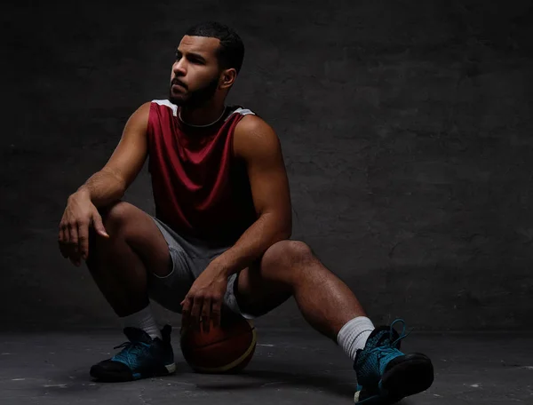 Junger afrikanisch-amerikanischer Basketballspieler in Sportbekleidung sitzt auf einem Ball vor dunklem Hintergrund. — Stockfoto