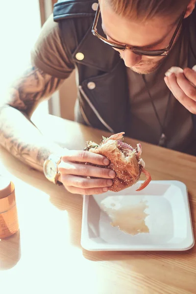 Portrait of a hungry hipster guy with a stylish haircut and beard sits at a table, decided to dine at a roadside cafe, eating a hamburger. — Stock Photo, Image