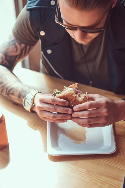Portrét chlapa hlad bokovky s stylový účes a plnovous sedí u stolu, rozhodl se najíst v silničních kavárna, jíst hamburger. — Stock fotografie