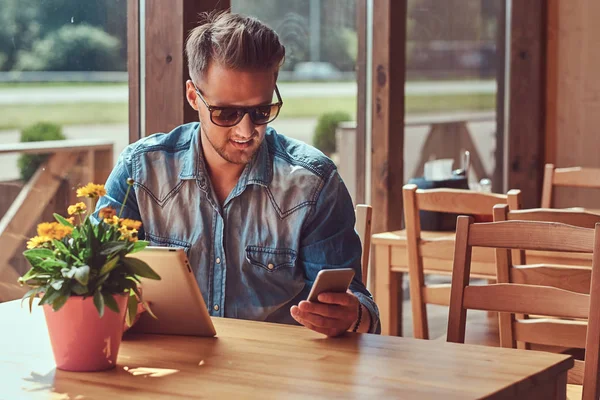 Hipster avec une coiffure élégante et la barbe est assis à une table dans un café en bord de route, ressemble quelque chose dans la tablette . — Photo