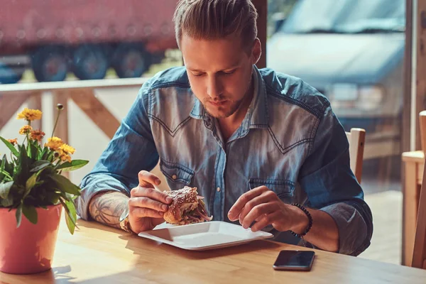 Portrait of a hungry hipster guy with a stylish haircut and beard sits at a table, decided to dine at a roadside cafe, eating a hamburger. — Stock Photo, Image
