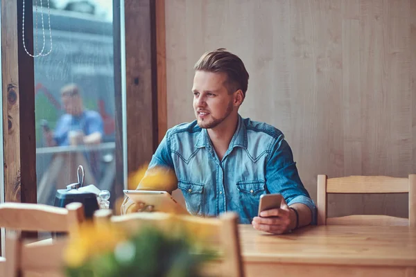 Hipster τύπος με ένα μοντέρνο κούρεμα και γενειάδα κάθεται σε ένα τραπέζι σε μια άκρη του δρόμου café, κατέχει ένα tablet και το smartphone. — Φωτογραφία Αρχείου