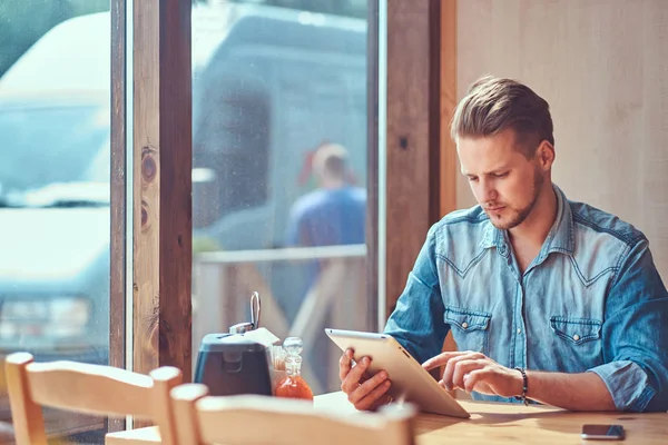 Hipster avec une coiffure élégante et la barbe est assis à une table dans un café en bord de route, ressemble quelque chose dans la tablette . — Photo