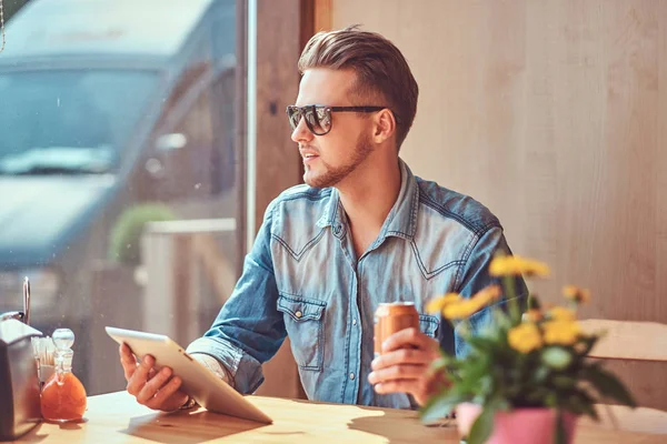 Hipster guy with a stylish haircut and beard sits at a table in a roadside cafe, holds soda and tablet computer while looking at the street. — Stock Photo, Image