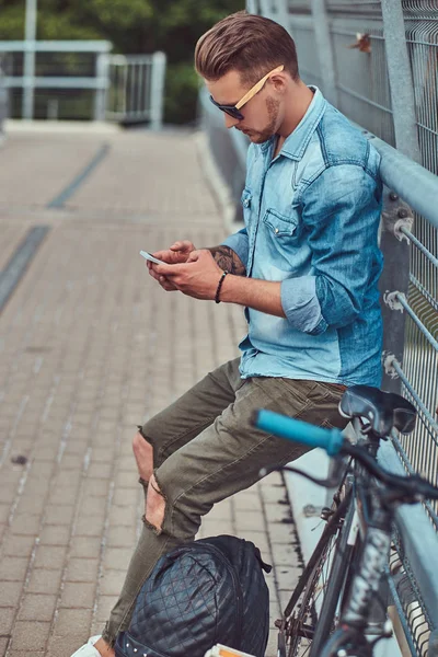 Красивий хіпстер зі стильною зачіскою в сонцезахисних окулярах відпочиває після їзди на велосипеді, використовуючи смартфон . — стокове фото