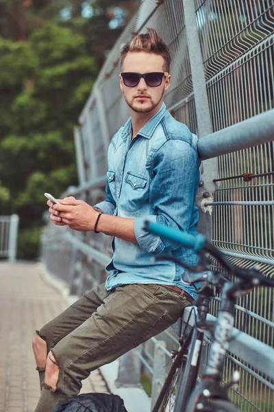Красивий хіпстер зі стильною зачіскою в сонцезахисних окулярах, відпочиваючи після їзди на велосипеді, тримає смартфон . — стокове фото