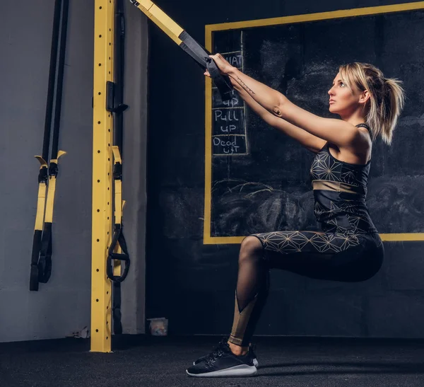 Sexy blonde woman in sportswear doing exercise with TRX system at gym. — Stock Photo, Image