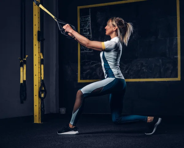 Sexy blonde woman in sportswear doing exercise with TRX system at gym. — Stock Photo, Image