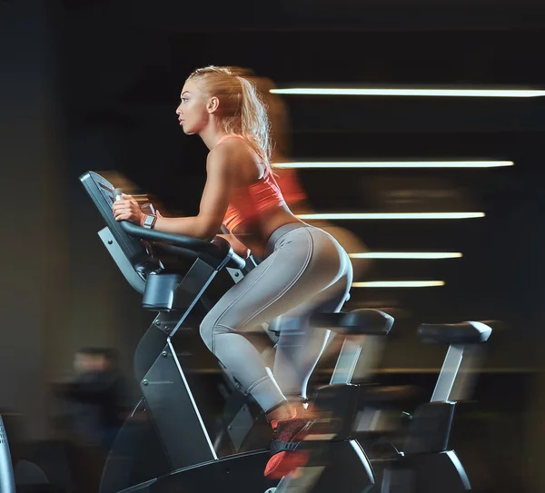 Sportive blonde woman working out on an exercise bike in modern fitness center. Fitness lifestyle in sport club. — Stock Photo, Image
