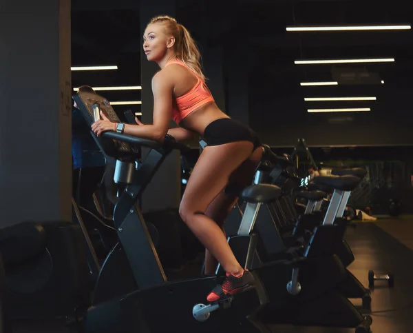 Молода красива блондинка в спортивному одязі тренується на тренажерному велосипеді в сучасному фітнес-центрі . — стокове фото