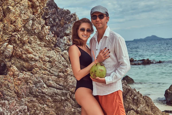 Casal atraente, segura coco com um tubo, de pé em uma praia perto de grandes pedras de recife, goza de umas férias em uma bela ilha . — Fotografia de Stock
