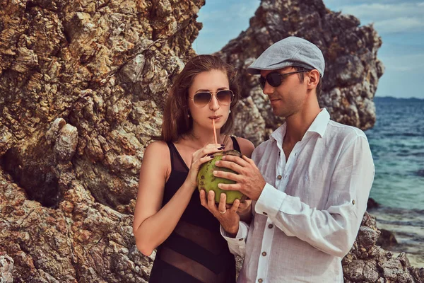 Casal atraente, segura coco com um tubo, de pé em uma praia perto de grandes pedras de recife, goza de umas férias em uma bela ilha . — Fotografia de Stock