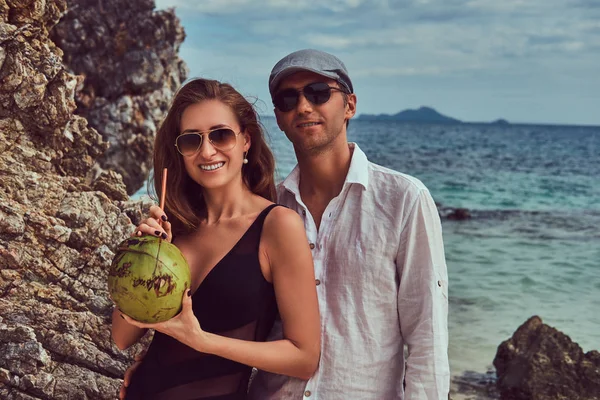 Coppia attraente, tiene cocco con un tubo, in piedi su una spiaggia vicino a grandi scogli, gode di una vacanza su una bella isola . — Foto Stock