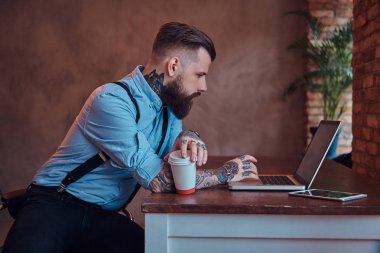 Bir dizüstü bilgisayarda çalışan yakışıklı dövmeli yenilikçi bir gömlek ve pantolon askısı masa başında oturan bir ofiste çatı iç paket servisi olan restoran kahve tutar.