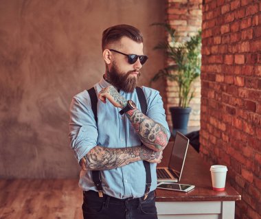 Çatı katı iç bir ofiste pencere bakarak eski moda dövmeli yenilikçi bir gömlek ve pantolon askısı, yakınındaki bir resepsiyon ile bir dizüstü bilgisayar, çapraz kolları olan daimi bir güneş gözlüğü giyiyor.