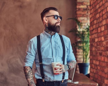 Çatı katı iç bir ofiste pencere bakarak eski moda dövmeli yenilikçi bir gömlek ve pantolon askısı, bir dizüstü bilgisayar ile bir Danışma yakınındaki bir kahve ile ayakta bir güneş gözlüğü giyiyor.