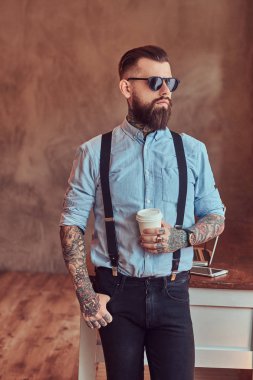 Çatı katı iç bir ofiste pencere bakarak eski moda dövmeli yenilikçi bir gömlek ve pantolon askısı, bir dizüstü bilgisayar ile bir Danışma yakınındaki bir kahve ile ayakta bir güneş gözlüğü giyiyor.