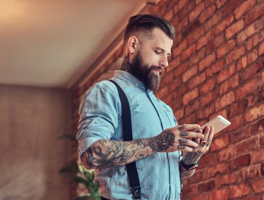 Bir çatı katı iç bir ofiste bir resepsiyon yakın dururken tablet kullanma bir gömlek ve pantolon askısı, bir güneş gözlüğü takan eski moda dövmeli hipster.